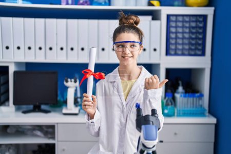 Foto de Adolescente chica trabajando en el laboratorio científico sosteniendo grado apuntando pulgar hacia el lado sonriendo feliz con la boca abierta - Imagen libre de derechos