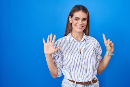 Foto de Mujer joven hispana de pie sobre fondo azul mostrando y señalando hacia arriba con los dedos número seis mientras sonríe confiada y feliz. - Imagen libre de derechos