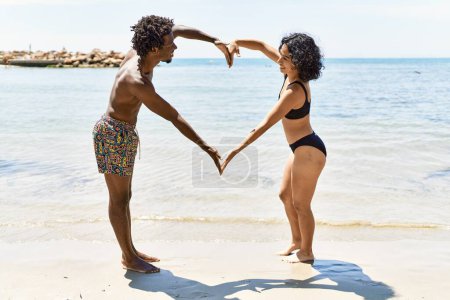 Téléchargez les photos : Jeune couple de touristes interracial portant des maillots de bain faisant grand cœur avec les bras à la plage. - en image libre de droit