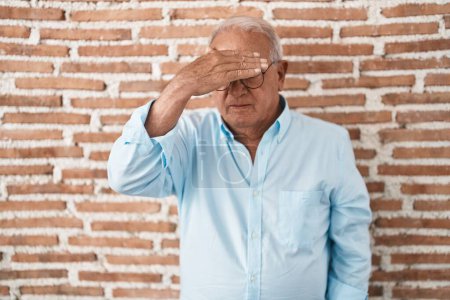 Photo for Senior man with grey hair standing over bricks wall covering eyes with hand, looking serious and sad. sightless, hiding and rejection concept - Royalty Free Image