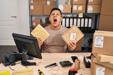 Foto de Joven hispano trabajando en el comercio electrónico de pequeñas empresas sosteniendo paquetes enojado y loco gritando frustrado y furioso, gritando con ira mirando hacia arriba. - Imagen libre de derechos