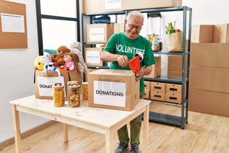 Photo for Senior man wearing volunteer uniform packing donations cardboard box at charity center - Royalty Free Image