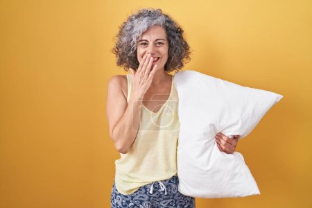 Téléchargez les photos : Femme d'âge moyen avec les cheveux gris portant un pyjama étreignant oreiller rire et rire embarrassé couvrant la bouche avec les mains, commérages et concept de scandale - en image libre de droit