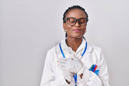 Téléchargez les photos : Femme africaine avec des tresses portant l'uniforme de scientifique souriant avec les mains sur la poitrine, les yeux fermés avec geste reconnaissant sur le visage. concept de santé. - en image libre de droit