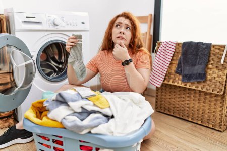 Photo for Young redhead woman putting dirty laundry into washing machine thinking worried about a question, concerned and nervous with hand on chin - Royalty Free Image