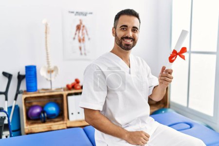 Photo for Young hispanic man wearing physiotherapist uniform holding diploma at clinic - Royalty Free Image