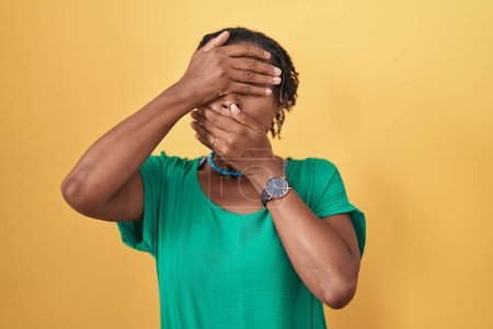 Foto de Mujer africana con rastas de pie sobre fondo amarillo cubriendo los ojos y la boca con las manos, sorprendida y conmocionada. ocultar emociones - Imagen libre de derechos