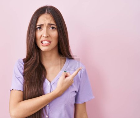 Photo for Young hispanic woman with long hair standing over pink background pointing aside worried and nervous with forefinger, concerned and surprised expression - Royalty Free Image