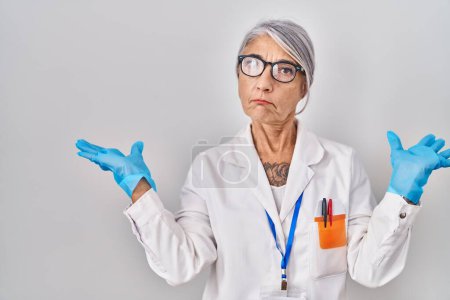 Photo for Middle age woman with grey hair wearing scientist robe clueless and confused expression with arms and hands raised. doubt concept. - Royalty Free Image