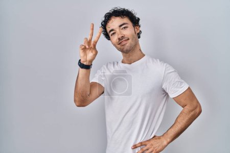 Téléchargez les photos : Homme hispanique debout sur fond isolé souriant en regardant la caméra montrant les doigts faisant signe de victoire. numéro deux. - en image libre de droit
