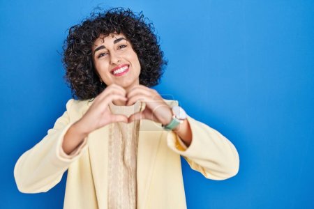Photo for Young brunette woman with curly hair standing over blue background smiling in love doing heart symbol shape with hands. romantic concept. - Royalty Free Image