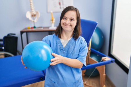 Photo for Down syndrome woman wearing physiotherapy uniform holding ball at physiotherapist clinic - Royalty Free Image