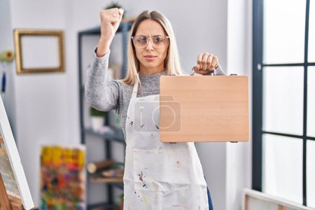 Photo for Young blonde painter woman holding wooden case at art studio annoyed and frustrated shouting with anger, yelling crazy with anger and hand raised - Royalty Free Image