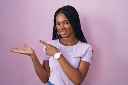 Foto de Mujer afroamericana con trenzas de pie sobre fondo rosa sorprendida y sonriendo a la cámara mientras se presenta con la mano y señalando con el dedo. - Imagen libre de derechos