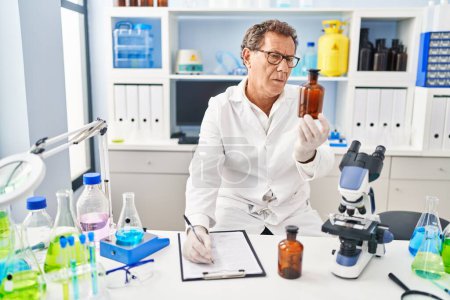 Téléchargez les photos : Middle age man wearing scientist uniform holding bottle writing on clipboard at laboratory - en image libre de droit