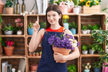 Téléchargez les photos : Jeune femme caucasienne travaillant au magasin de fleuriste tenant pot avec des fleurs surpris par une idée ou une question pointant du doigt avec le visage heureux, numéro un - en image libre de droit