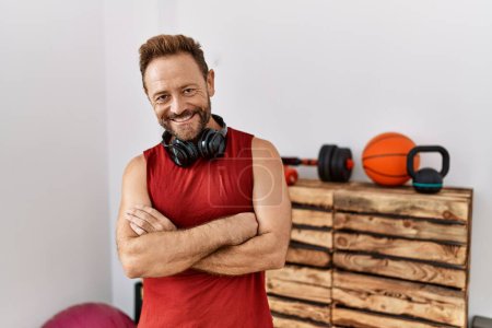 Téléchargez les photos : Homme d'âge moyen portant des vêtements de sport et des écouteurs à la salle de gym visage heureux souriant avec les bras croisés regardant la caméra. personne positive. - en image libre de droit