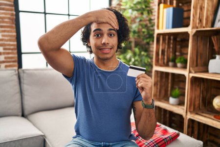 Téléchargez les photos : Homme hispanique avec les cheveux bouclés tenant la carte de crédit stressé et frustré avec la main sur la tête, visage surpris et en colère - en image libre de droit