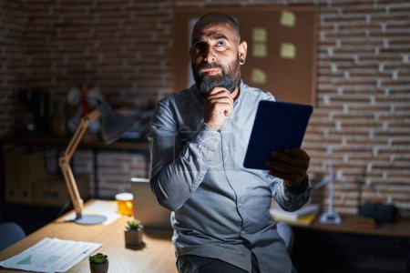 Photo for Young hispanic man with beard and tattoos working at the office at night thinking concentrated about doubt with finger on chin and looking up wondering - Royalty Free Image