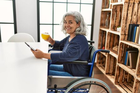 Foto de Mujer discapacitada de pelo gris de mediana edad usando touchpad y cofffee para beber sentada en silla de ruedas en casa. - Imagen libre de derechos