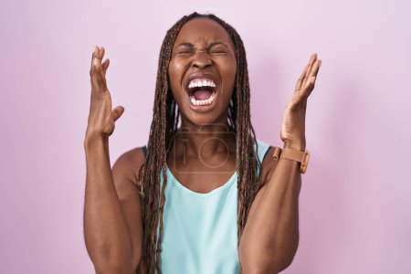 Foto de Mujer afroamericana de pie sobre fondo rosa loca y loca gritando y gritando con expresión agresiva y los brazos levantados. concepto de frustración. - Imagen libre de derechos