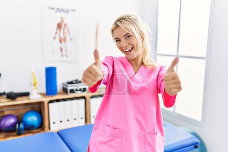 Photo for Young caucasian woman working at pain recovery clinic approving doing positive gesture with hand, thumbs up smiling and happy for success. winner gesture. - Royalty Free Image