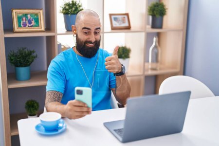 Foto de Young hispanic man with beard and tattoos doing video call with smartphone smiling happy and positive, thumb up doing excellent and approval sign - Imagen libre de derechos
