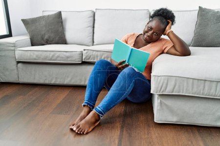 Foto de Young african american woman reading book sitting on the floor at home - Imagen libre de derechos