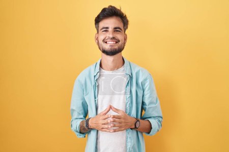 Téléchargez les photos : Jeune homme hispanique avec des tatouages debout sur fond jaune mains ensemble et doigts croisés souriant détendu et joyeux. succès et optimisme - en image libre de droit