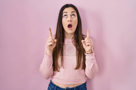 Photo for Young brunette woman standing over pink background amazed and surprised looking up and pointing with fingers and raised arms. - Royalty Free Image
