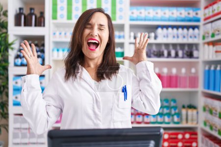 Foto de Middle age brunette woman working at pharmacy drugstore celebrating mad and crazy for success with arms raised and closed eyes screaming excited. winner concept - Imagen libre de derechos