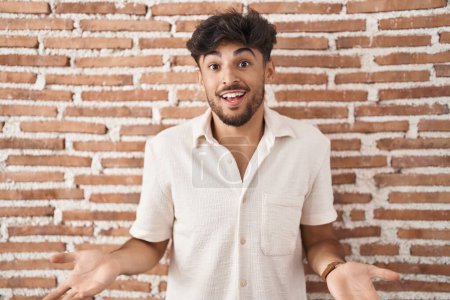 Photo for Arab man with beard standing over bricks wall background smiling cheerful with open arms as friendly welcome, positive and confident greetings - Royalty Free Image