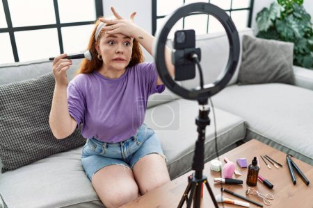 Téléchargez les photos : Jeune rousse femme enregistrement maquillage tutoriel avec smartphone à la maison stressé et frustré avec la main sur la tête, visage surpris et en colère - en image libre de droit