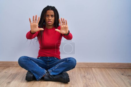 Téléchargez les photos : Jeune afro-américain avec des tresses assises sur le sol à la maison faisant geste d'arrêt avec les mains paumes de main, expression de colère et de frustration - en image libre de droit