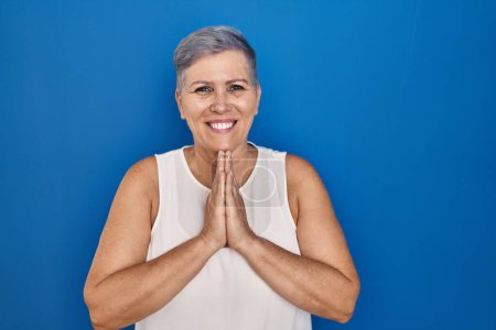 Foto de Mujer caucásica de mediana edad de pie sobre fondo azul rezando con las manos juntas pidiendo perdón sonriendo confiado. - Imagen libre de derechos