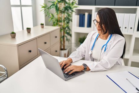 Photo for Young hispanic woman wearing doctor uniform using laptop working at clinic - Royalty Free Image