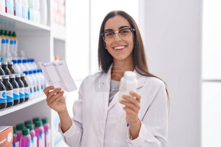 Téléchargez les photos : Jeune belle hispanique femme pharmacien tenant des pilules bouteille lecture prescription à la pharmacie - en image libre de droit