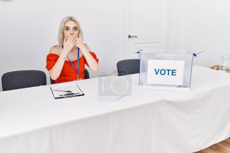 Foto de Joven mujer caucásica en las elecciones políticas sentada por votación impactada cubriendo la boca con las manos por error. concepto secreto. - Imagen libre de derechos