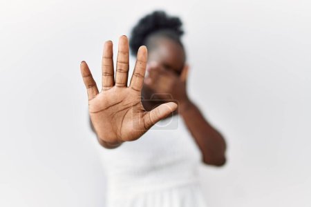 Téléchargez les photos : Jeune femme africaine debout sur fond blanc isolé couvrant les yeux des mains et faisant un geste d'arrêt avec expression triste et peur. concept embarrassé et négatif. - en image libre de droit