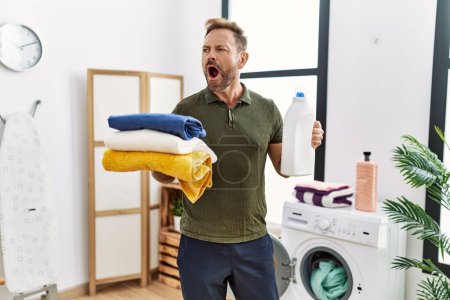 Foto de Hombre de mediana edad sosteniendo la ropa limpia y la botella de detergente enojado y loco gritando frustrado y furioso, gritando con ira. rabia y concepto agresivo. - Imagen libre de derechos