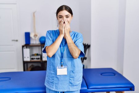 Photo for Young hispanic woman wearing physiotherapist uniform standing at clinic laughing and embarrassed giggle covering mouth with hands, gossip and scandal concept - Royalty Free Image