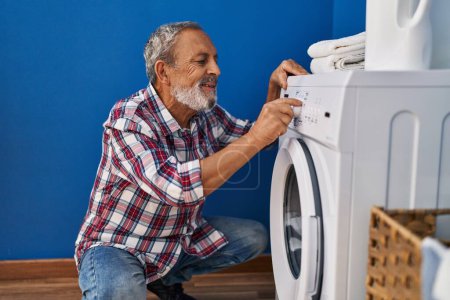Foto de Hombre mayor de pelo gris sonriendo confiado encendiendo la lavadora en la lavandería - Imagen libre de derechos