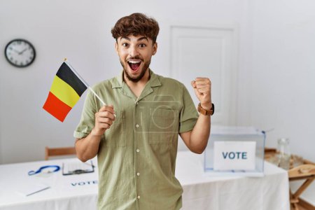 Foto de Young arab man at political campaign election holding belgium flag screaming proud, celebrating victory and success very excited with raised arms - Imagen libre de derechos