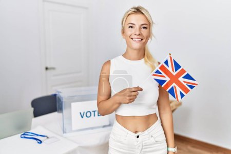 Téléchargez les photos : Jeune femme caucasienne à l'élection de campagne politique tenant drapeau britannique regardant positif et heureux debout et souriant avec un sourire confiant montrant les dents - en image libre de droit