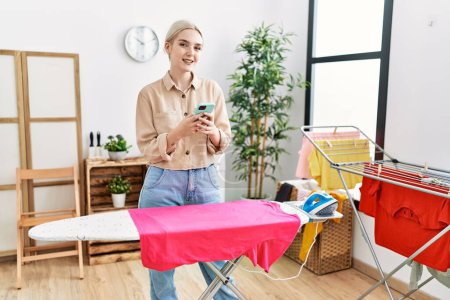 Téléchargez les photos : Young caucasian woman using smartphone ironing clothes at laundry room - en image libre de droit