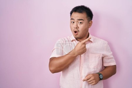 Foto de Chinese young man standing over pink background surprised pointing with finger to the side, open mouth amazed expression. - Imagen libre de derechos