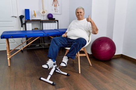 Photo for Senior caucasian man at physiotherapy clinic using pedal exerciser doing happy thumbs up gesture with hand. approving expression looking at the camera showing success. - Royalty Free Image