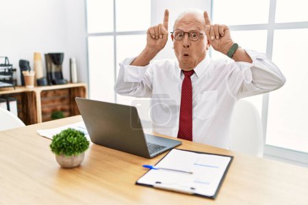 Téléchargez les photos : Senior homme travaillant au bureau en utilisant ordinateur portable faisant geste drôle avec doigt sur la tête comme cornes de taureau - en image libre de droit