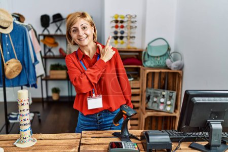 Photo for Middle age blonde woman working working as manager at retail boutique cheerful with a smile of face pointing with hand and finger up to the side with happy and natural expression on face - Royalty Free Image