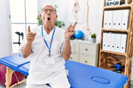 Foto de Hombre de fisioterapia senior que trabaja en la clínica de recuperación del dolor asombrado y sorprendido mirando hacia arriba y señalando con los dedos y los brazos elevados. - Imagen libre de derechos